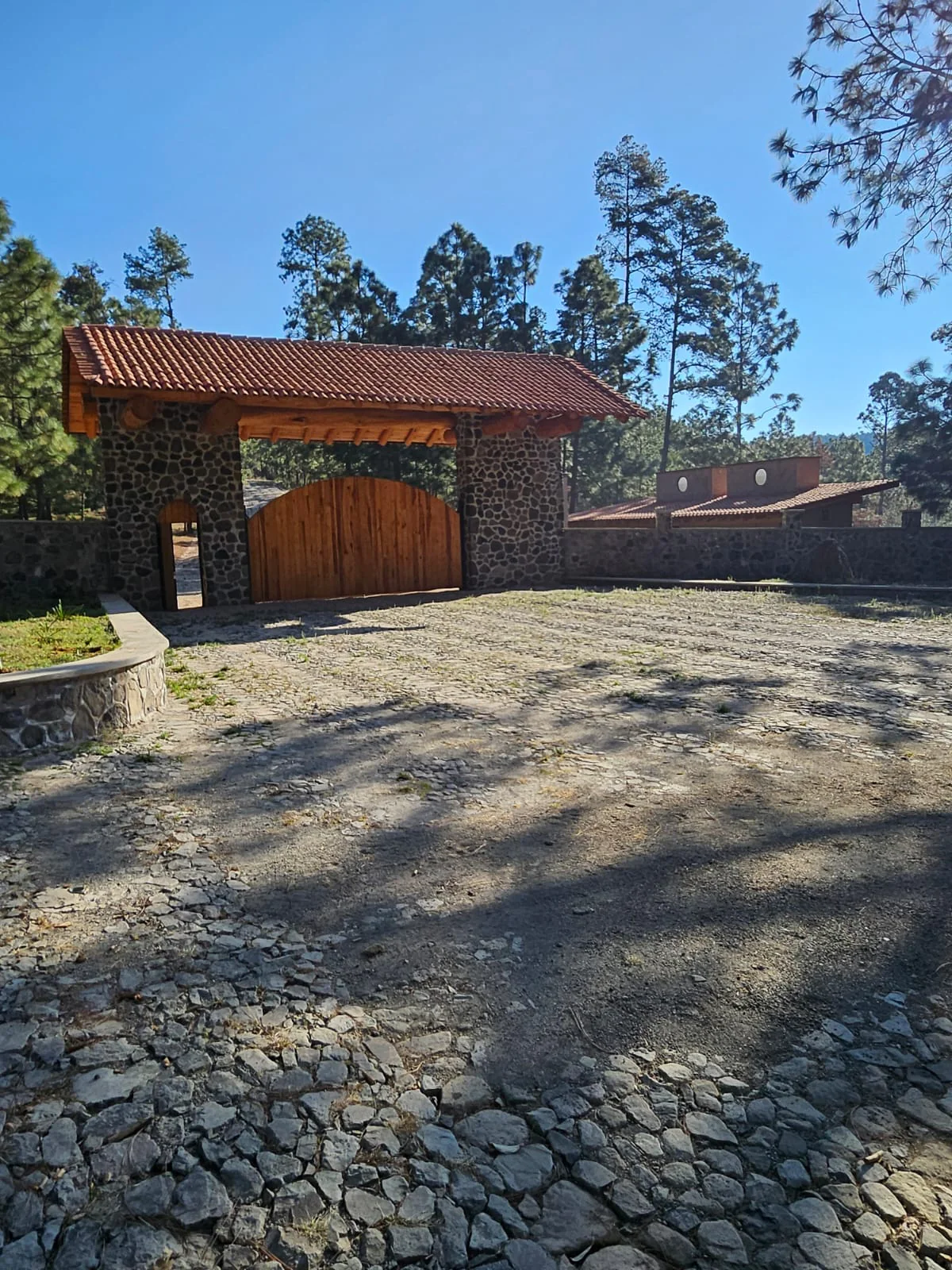 Ingreso de Tlalli Residencial con portón de madera y muro de piedra, rodeado de pinos en Tapalpa