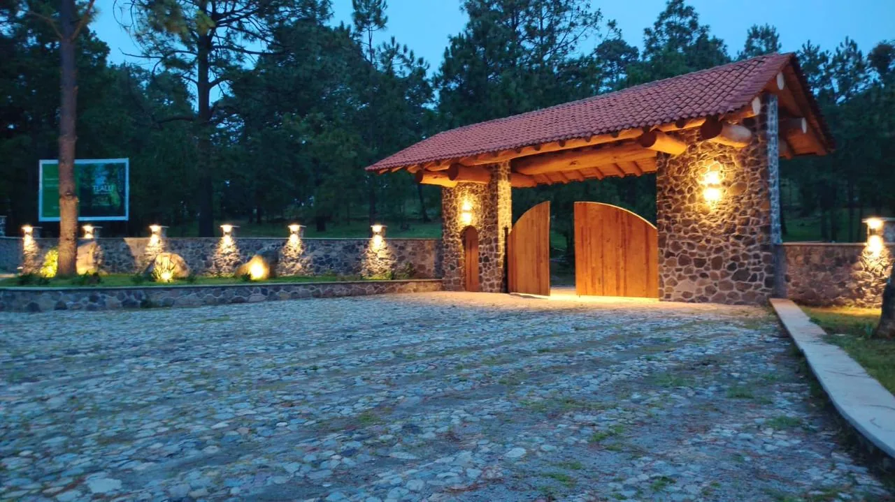 Ingreso de Tlalli Residencial con el portón de madera abierto, muro de piedra y bosque de pinos en Tapalpa